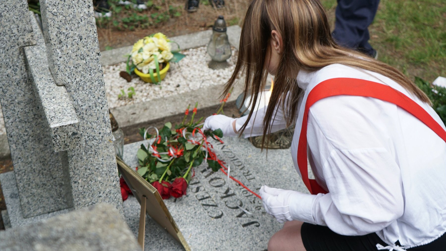 Zdjęcie przedstawia dziewczynę kładąca kwiaty na grobie.