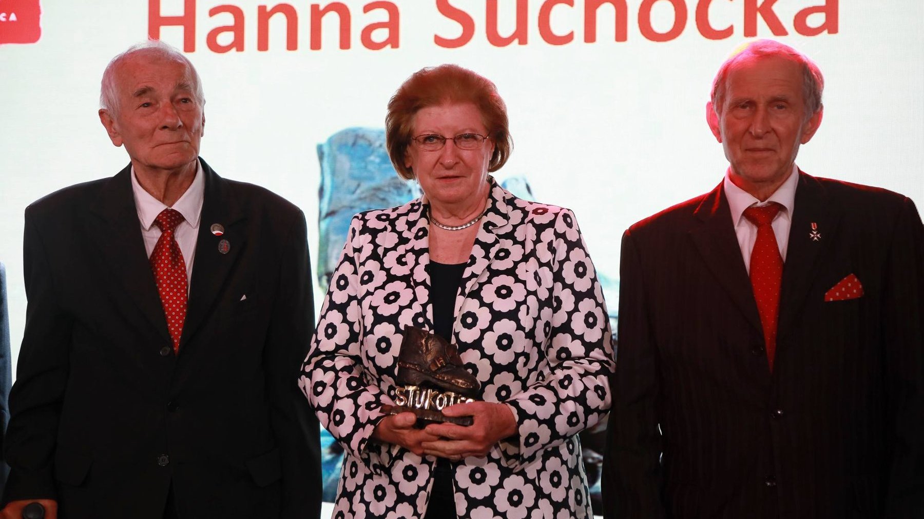 Na zdjęciu od lewej: Jerzy Majrzchak, Hanna Suchocka i Andrzej Sporny. Hanna Suchocka trzyma w rękach nagrodę Stukot'56. Mężczyźni sa ubrani w czarne garnitury, białe koszule i czerwone krawaty. Kobieta jest ubrana w czarną suknie i czarną marynarkę z printem białych kwiatów.