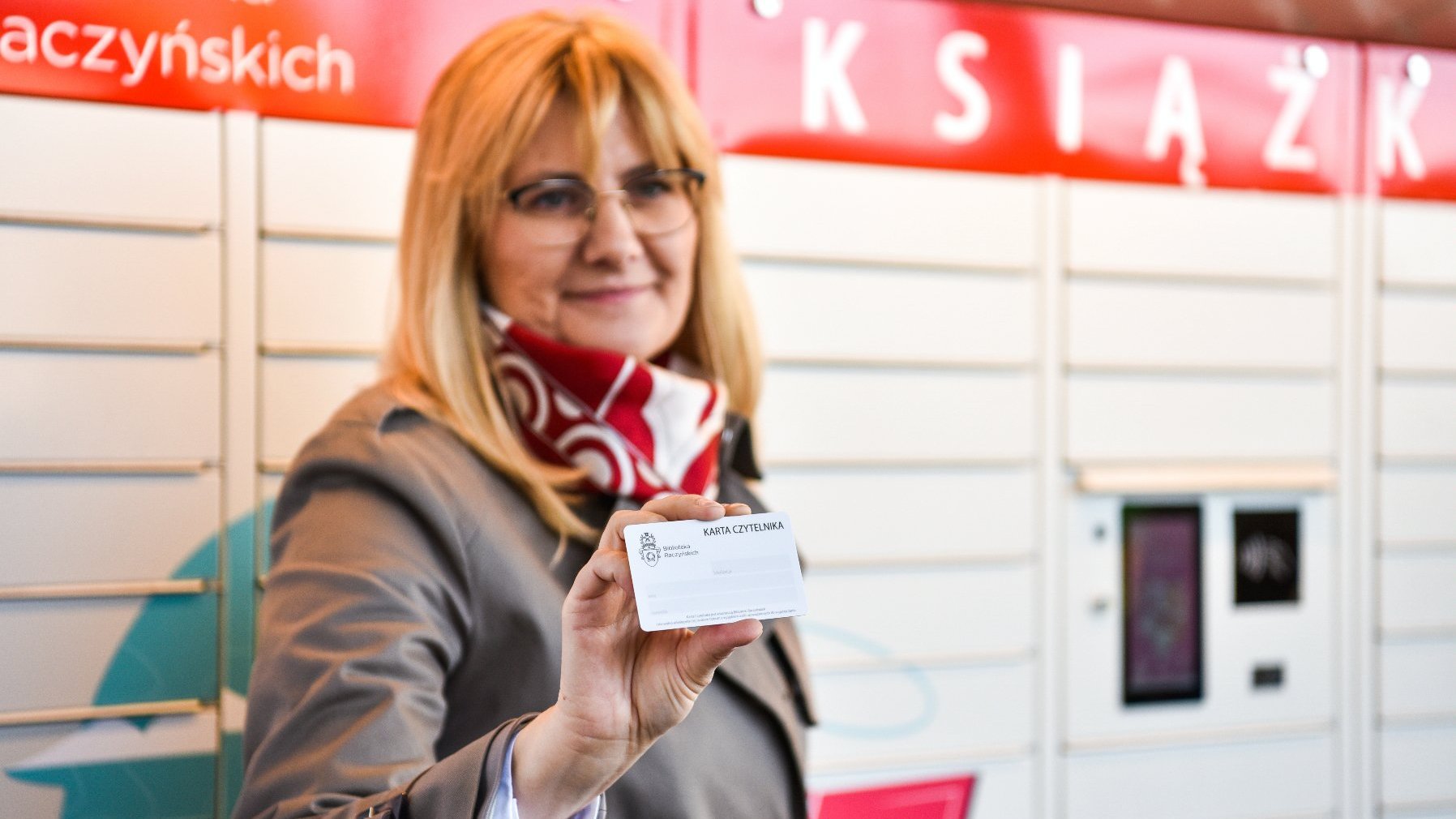 Galeria zdjęć przedstawia książkomat oraz stojących w jego sąsiedztwie ludzi, w tym dyrektorkę biblioteki, która prezentuje sposób obsługi urządzenia.