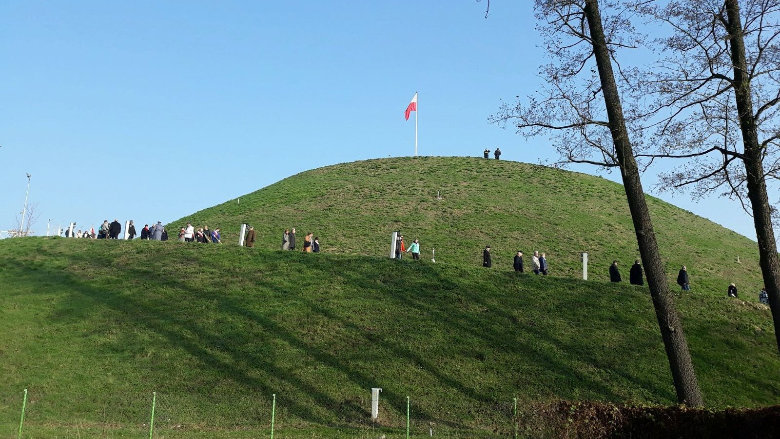 Popołudnie. Na szczycie Kopca stoi wysoki maszt z flagą Polski. Wokół Kopca chodzą ludzie, którzy wchodzą na szczyt lub schodzą na dół. W tle bezchmurne niebo. W kadrze po prawej są dwa drzewa.