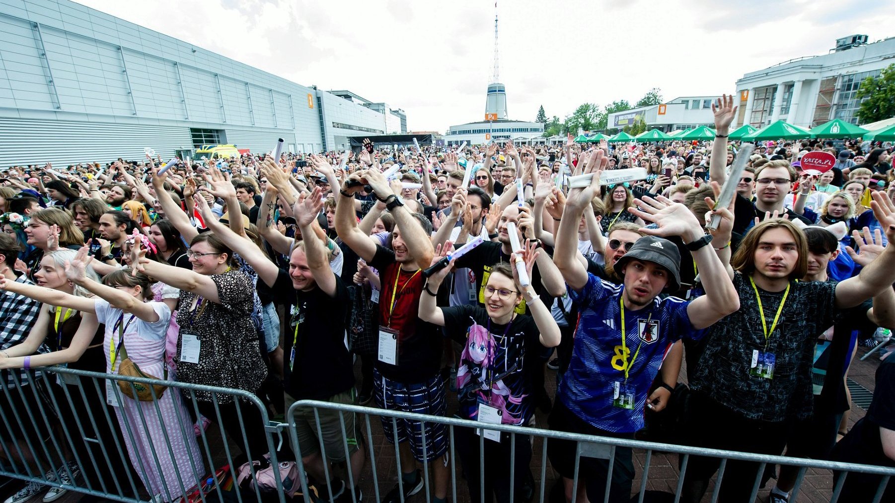 Widok ze sceny na tłum uczestników, którzy bawią się pod sceną koncertową. Uczestnicy mają podniesione ręce do góry, uśmiechają się, śpiewają, skaczą. W tle iglica Międzynarodowych Targów Poznańskich oraz inne pawilony.