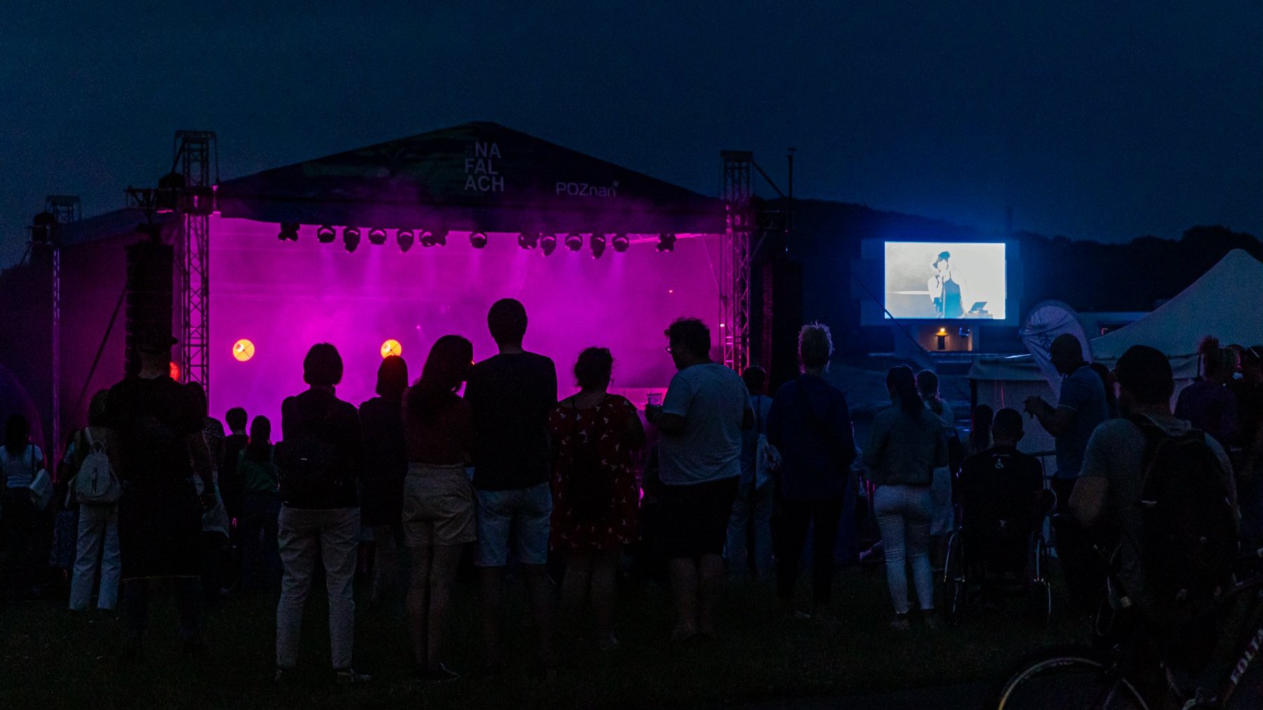 Zdjęcie zrobione nocą. Na pierwszym planie widownia, która stoi przodem do sceny. Przed nimi stoi scena, która jest oświetlona na różowo. Po prawej stronie stoi ekran, na którym wyświetla się osoba występująca na scenie.