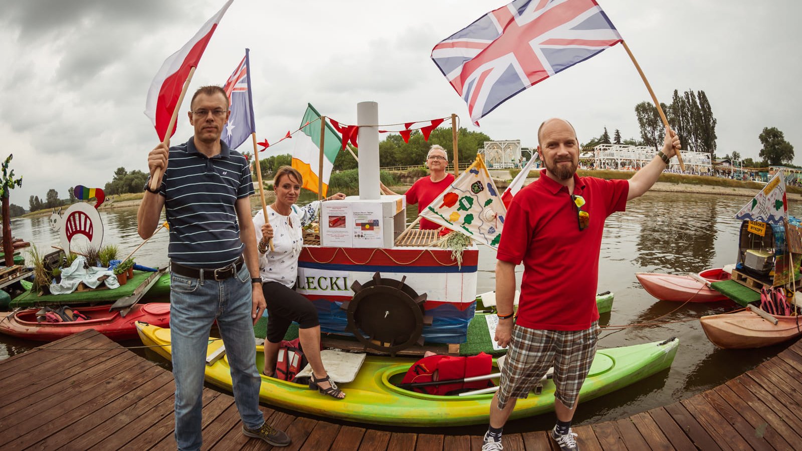 Trzech mężczyzn i kobieta pozują przy swojej intalacji. Na dwóch kajakach stoi mały statek z napisem "Strzelecki". Osoby trzymają flagę Polski, Wielkiej Brytanii, Irlandii i Australii. Obok nich stoją jeszcze dwie instalacje z osiedli.