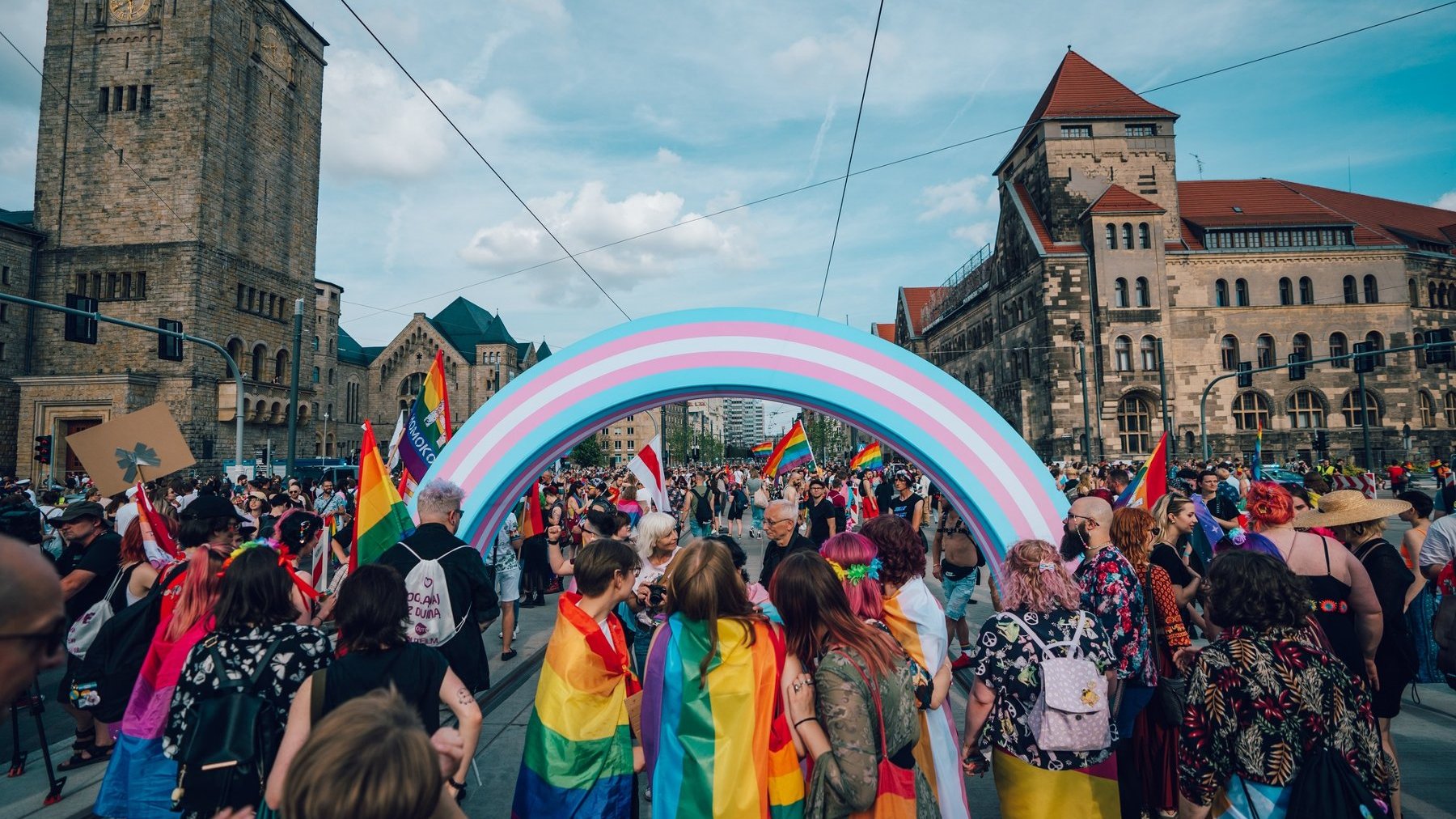 Zdjęcie z perspektywy osoby, która idzie w Marszu. Na pierwszym planie osoby odwrócone tyłem przykryte tęczowymi flagami. Przechodzą pod niebiesko-biało-różową tęczą, która reprezentuje grupę LGBt+. Wokół przechodzą inni ludzie trzymając w rękach kolorwe flagi i transparenty. Po lewej wieża CK Zamku, a po prawej budynek Filharmonii Poznańskiej.