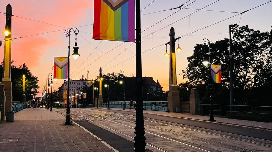 Zachód słońca. Widok na Most Teatralny w kierunku Jeżyc. Na latarniach powieszone są flagi, które reprezentują społeczność LGBT+. Są to flagi, które składają się z kolorowych pasków, z których każdy reprezentuje inną reprezentację LGBT+.