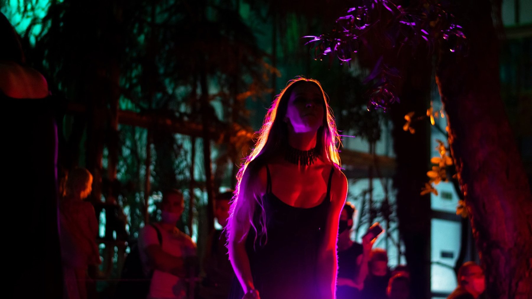 Młoda kobieta w czarnej sukience i w długich włosach jest oświetlona od dołu przez kolorowe neony. Nie widać jej twarzy, która jest skierowana w górę. Za kobietą kilka sylwetek innych gości.