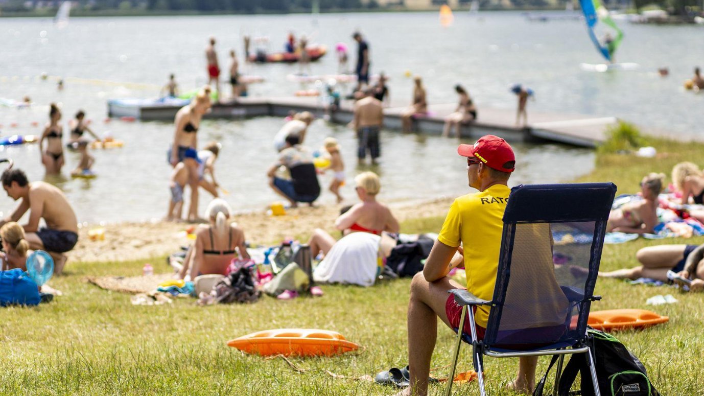 Na pierwszym planie mężczyzna siedzi na krześle letniskowym. Jest ubrany w żółtą koszulkę z napisem "Ratownik", czerwoną czapkę i kąpielówki. Obserwuje, co dzieje się w jeziorze. Wokół niego ludzie leżą na plaży lub kąpią się w jeziorze.
