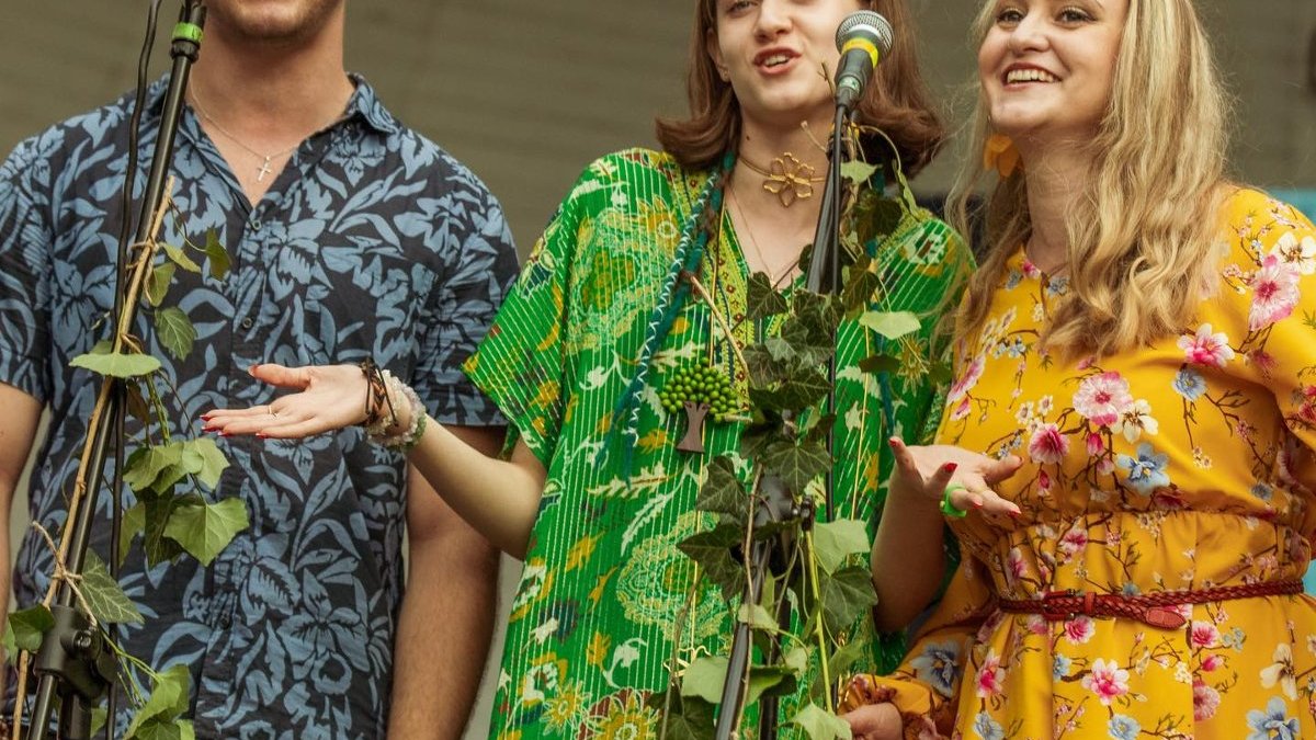 Mężczyna i dwie kobiety stoją obok siebie na scenie i śpiewają do mikrofonów. Są ubrani w kolorowe, kwiatowe stroje. Stojaki od mikrofonów są porośnięte liśćmi.