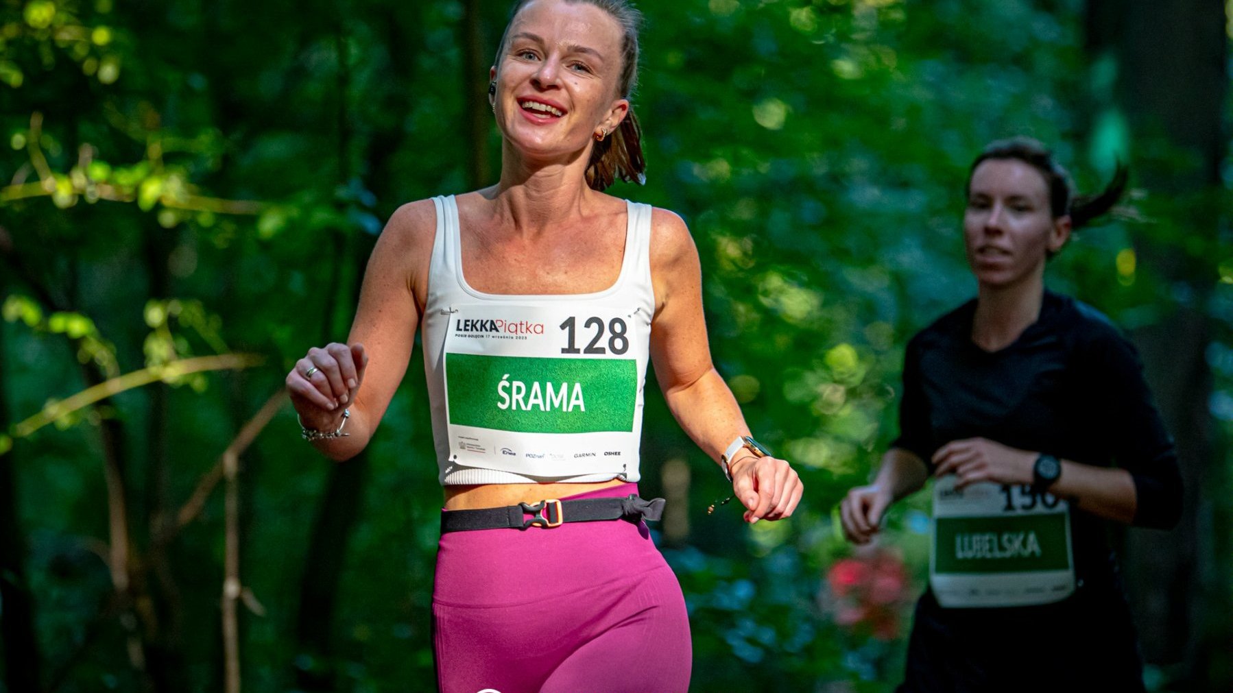 Dwie kobiety ubrane w stroje sportowe i z przyklejonymi numerami biegną w lesie.