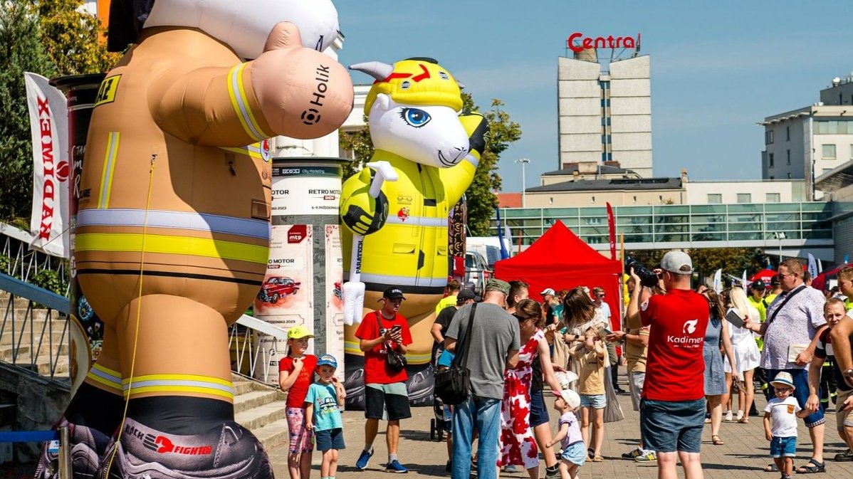 Pyrek i Tyrek, czyli symbol Poznania, jako zmuchane balony w strojach strażacki. Pod nimi dzieci pozują do zdjęć. Obok ustawia się kolejka do wejścia.