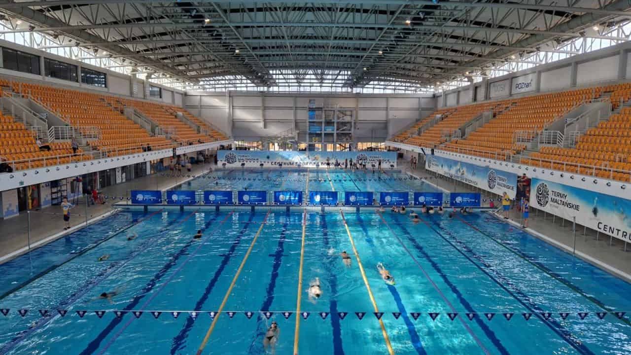 Hala basenowa Term Maltańskich. W centrum dziesięc torów pływackich na basenie sportowym. W wodzie pływają ludzie. Po lewej i po prawej trybuny z żółtymi krzesłami.