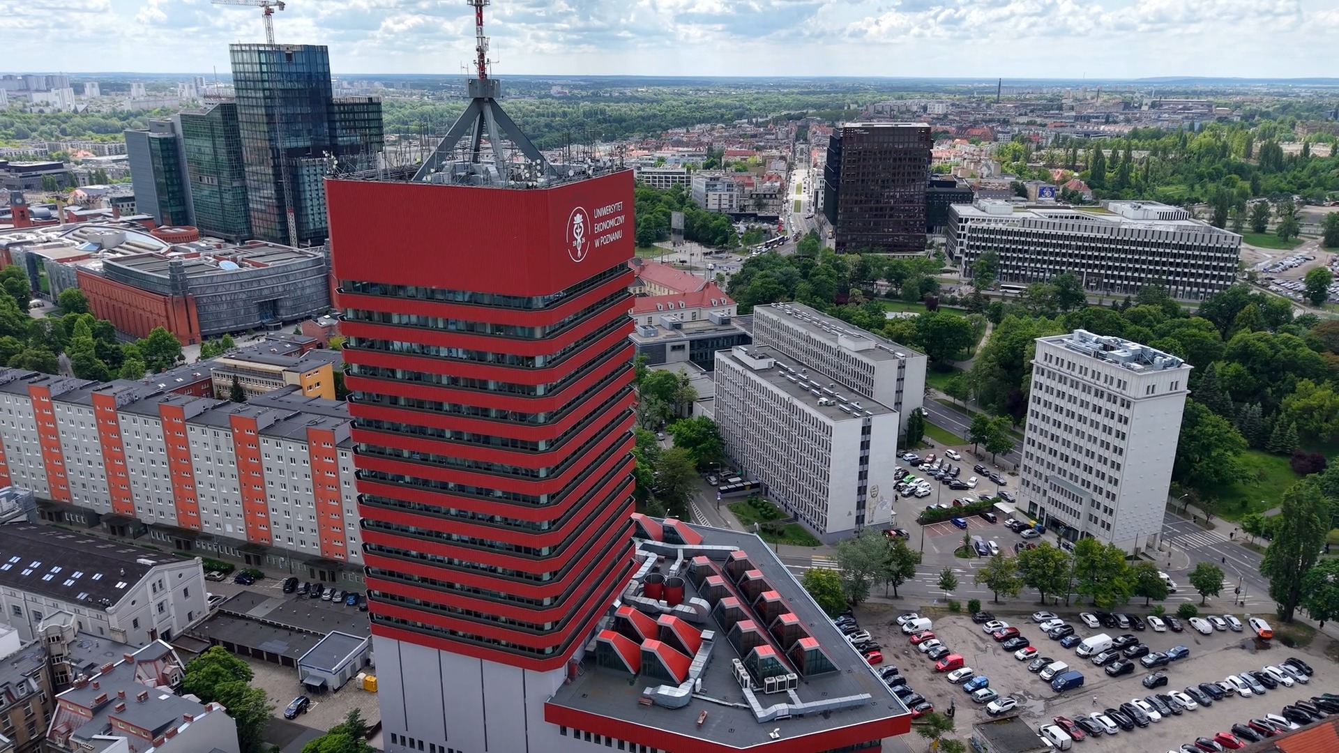 Widok na panoramę miasta? W samym centrum Poznania otwarto taras widokowy. Widok z lotu ptaka na czerwoną wieżę Uniwersytetu Ekonomicznego. Wokół inne budynki w okolicy, zielone drzewa. W tle widok na panoramę Poznania.