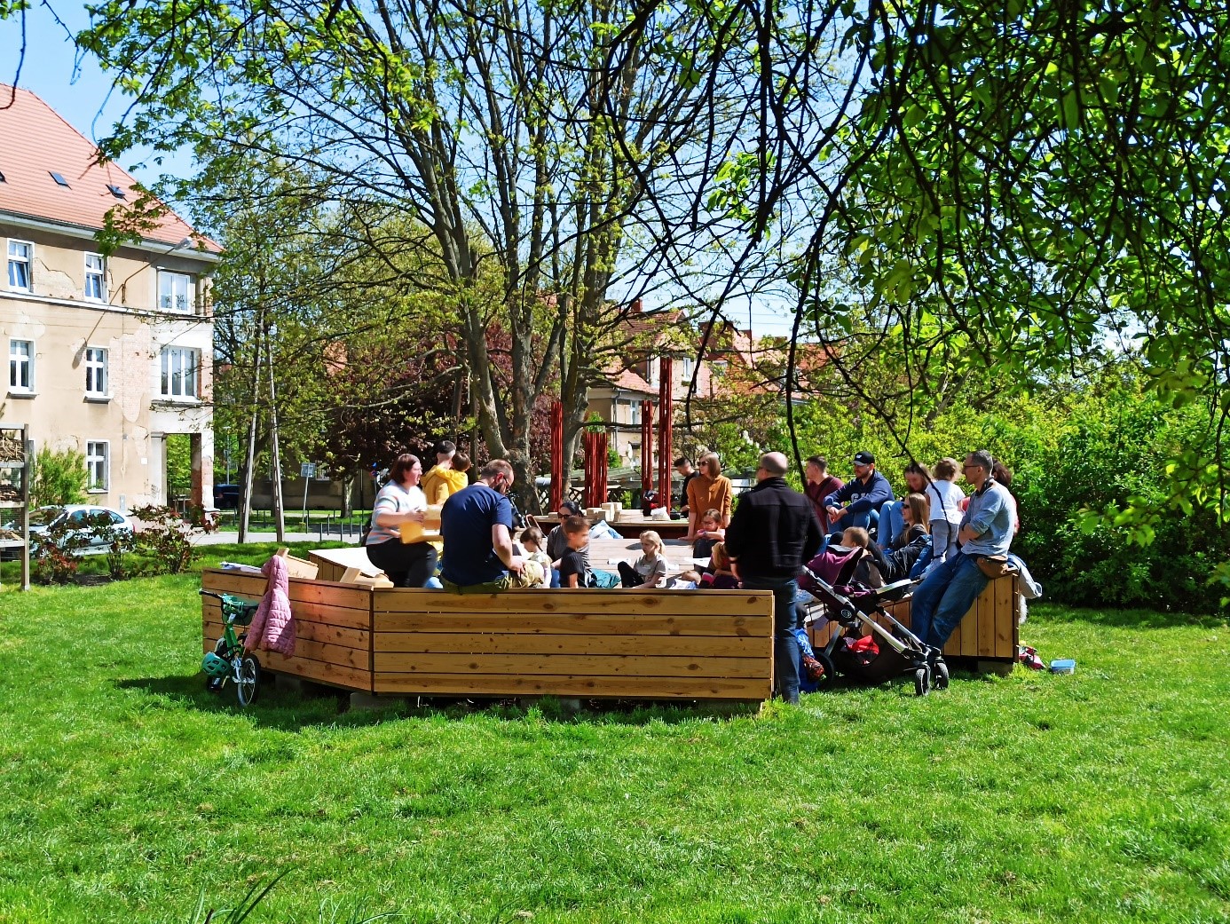 Grupa osób siedzących na ławkach wśród zielenii, w tle widać zabudowania wielorodzinne - grafika artykułu