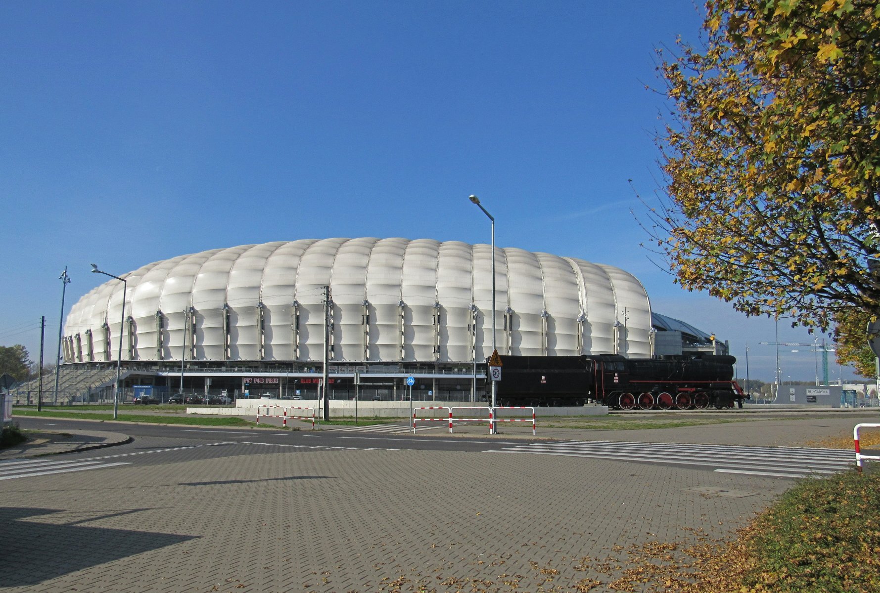 Zdjęcie przedstawia białą kopułę stadionu przy ul. Bułgarskiej. Przed nim stoi parowóz. - grafika artykułu