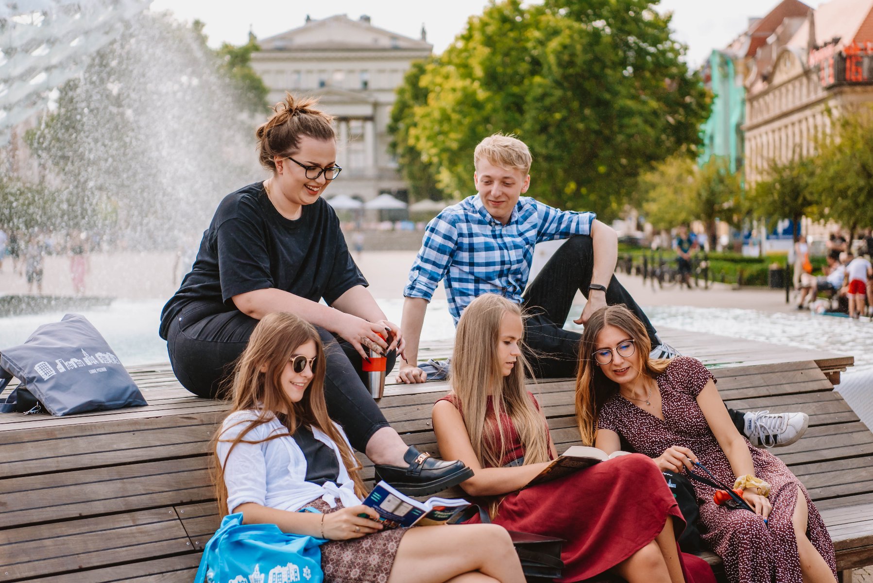 Na zdjęciu grupa młodych ludzi siedzi przy fontannie, część z nich ma w rękach książki - grafika artykułu