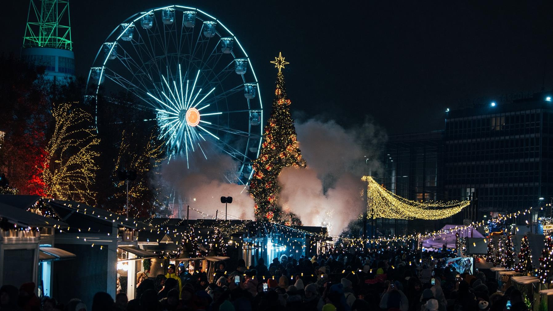 Jarmark na targach nocą. Na pierwszym planie grupa ludzi. W tle koło młyńskie i choinka.