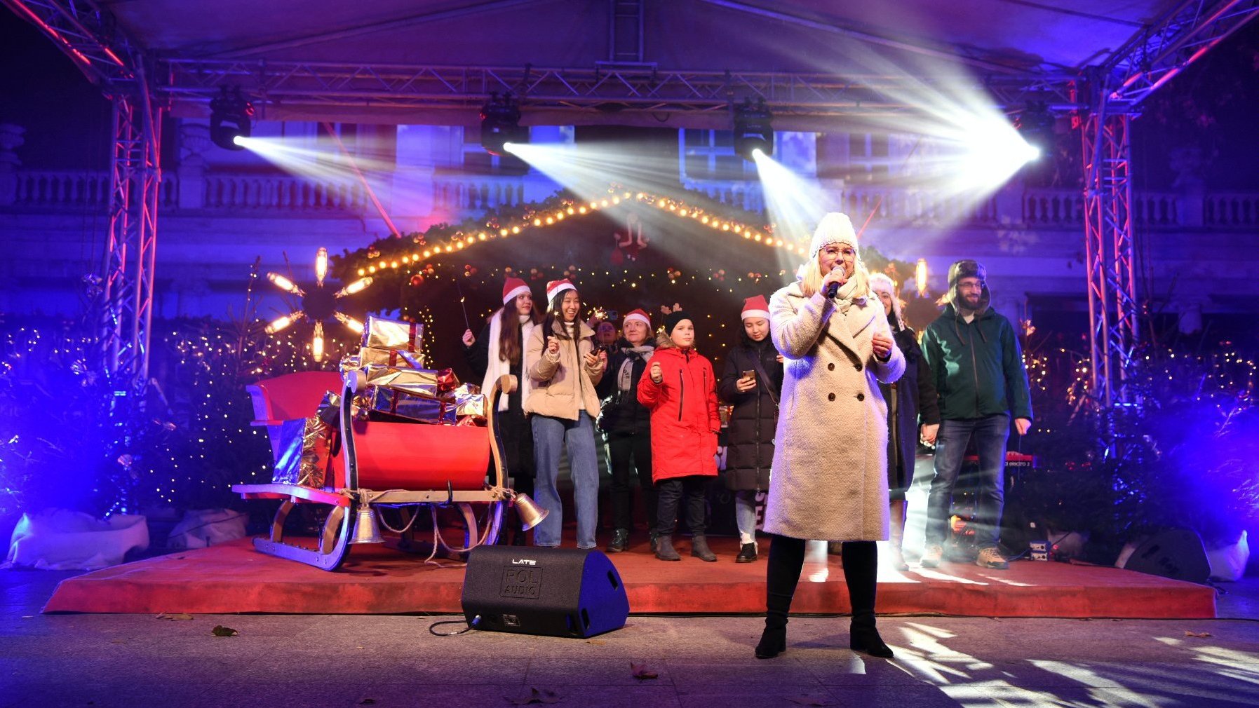 Grupa osób stoi na scenie przed budnkiem Arkadii. Jedna z nich - na pierwszym planie - w dłoni trzyma mikrofon, śpiewa.