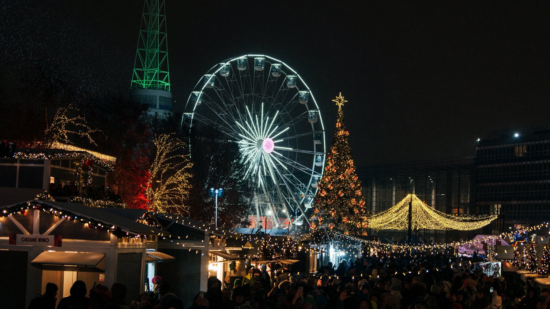 Zdjęcie przedstawia młyńskie koło na targach. Obok widać tłum ludzi, choinkę i iluminacje świąteczne.