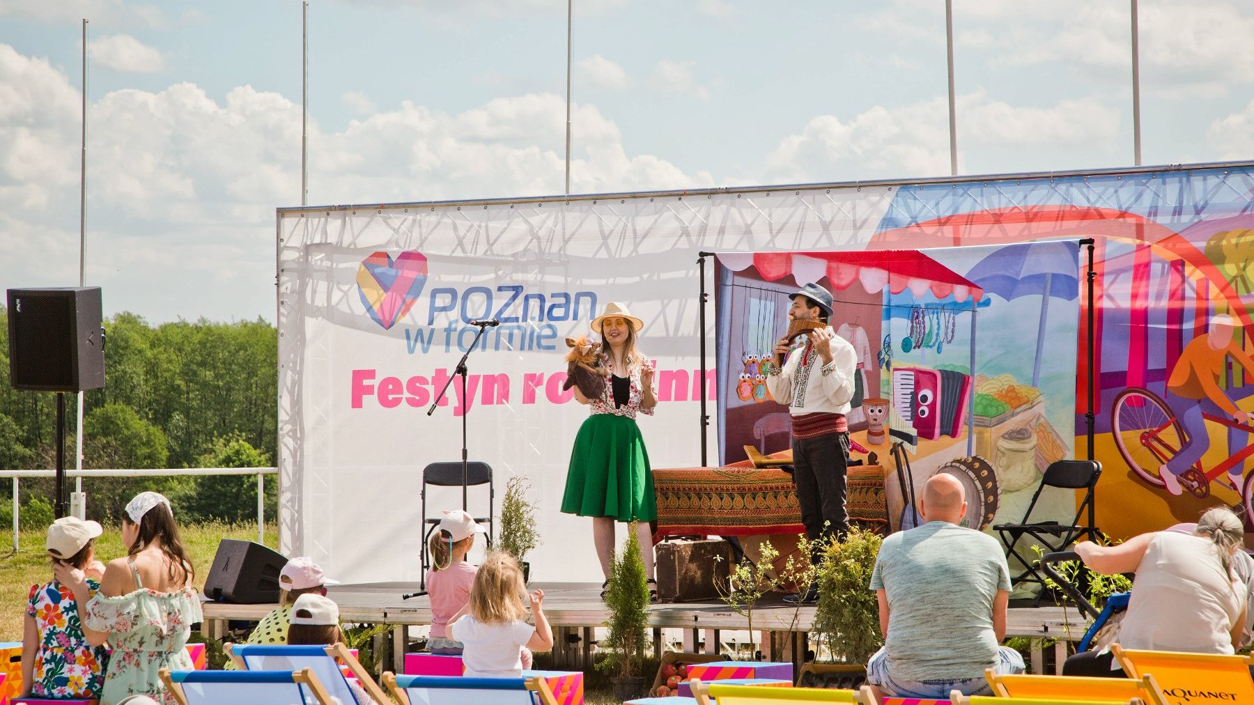 Na zdjęciu dzieci i dorośli przed sceną, a na scenie dwoje aktorów w kolorowych ubraniach