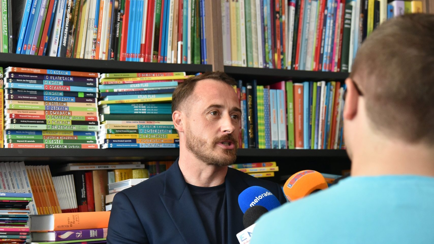 Zdjęcie przedstawia mężczyznę mówiącego do mikrofonu. Za jego plecami znajduje się półka z książkami.