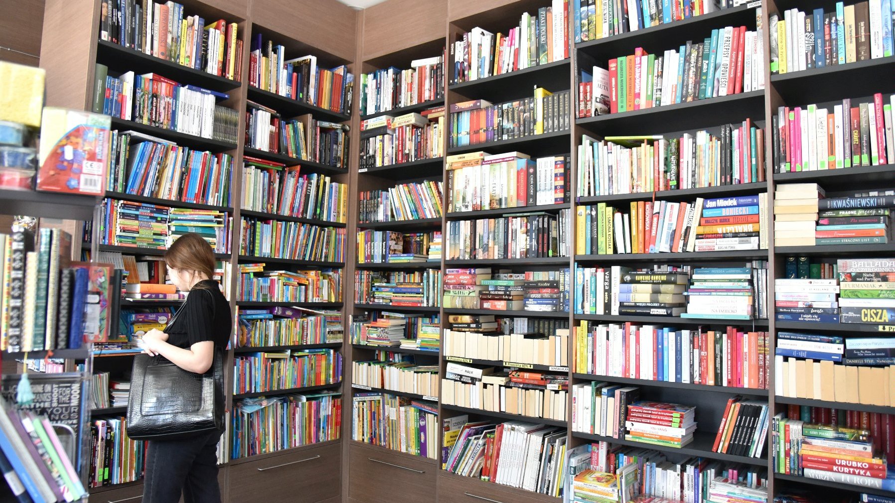 Zdjęcie przedstawia dziewczynę oglądającą książki stojące na półce.