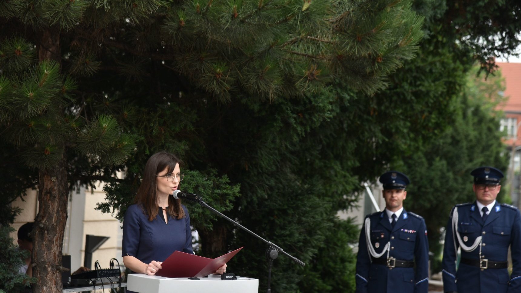 Na zdjęciu kobieta przy mikrofonie, obok dwóch policjantów