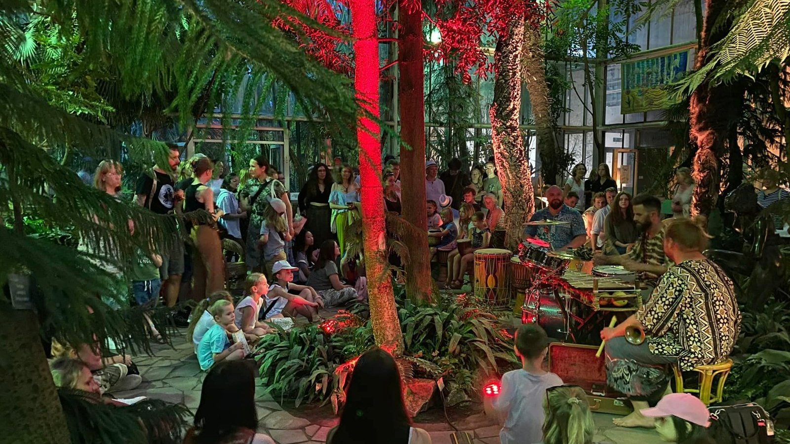Zdjęcie przedstawia tłum ludzi w palmiarni. Widać na nim egzotyczne rośliny oraz ludzi grających na różnych instrumentach.