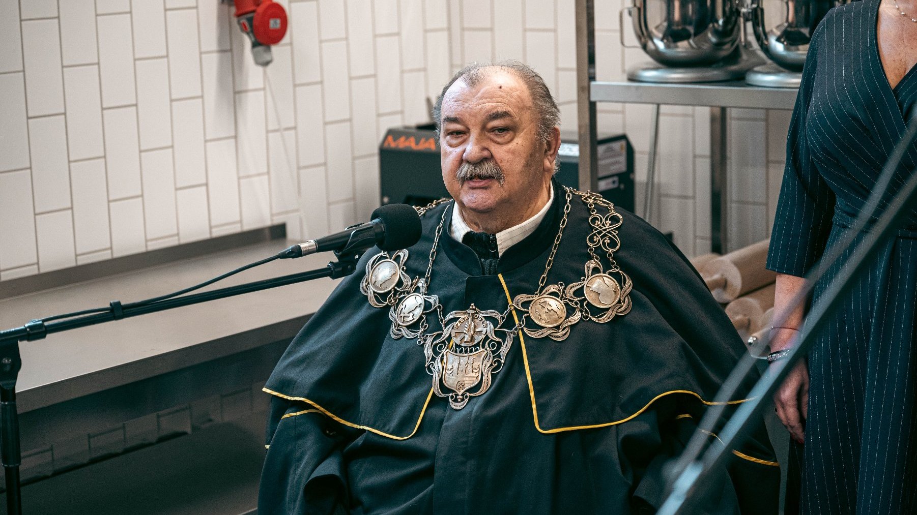 Osoba siedząca w kuchni lub w obszarze przygotowania żywności, ubrana w ceremonialny strój z dużym, ozdobnym łańcuchem na szyi, zawierającym kilka medalionów i centralny emblemat. Przed osobą znajduje się mikrofon, sugerując, że osoba może wygłaszać przemówienie lub być wywiadowana.