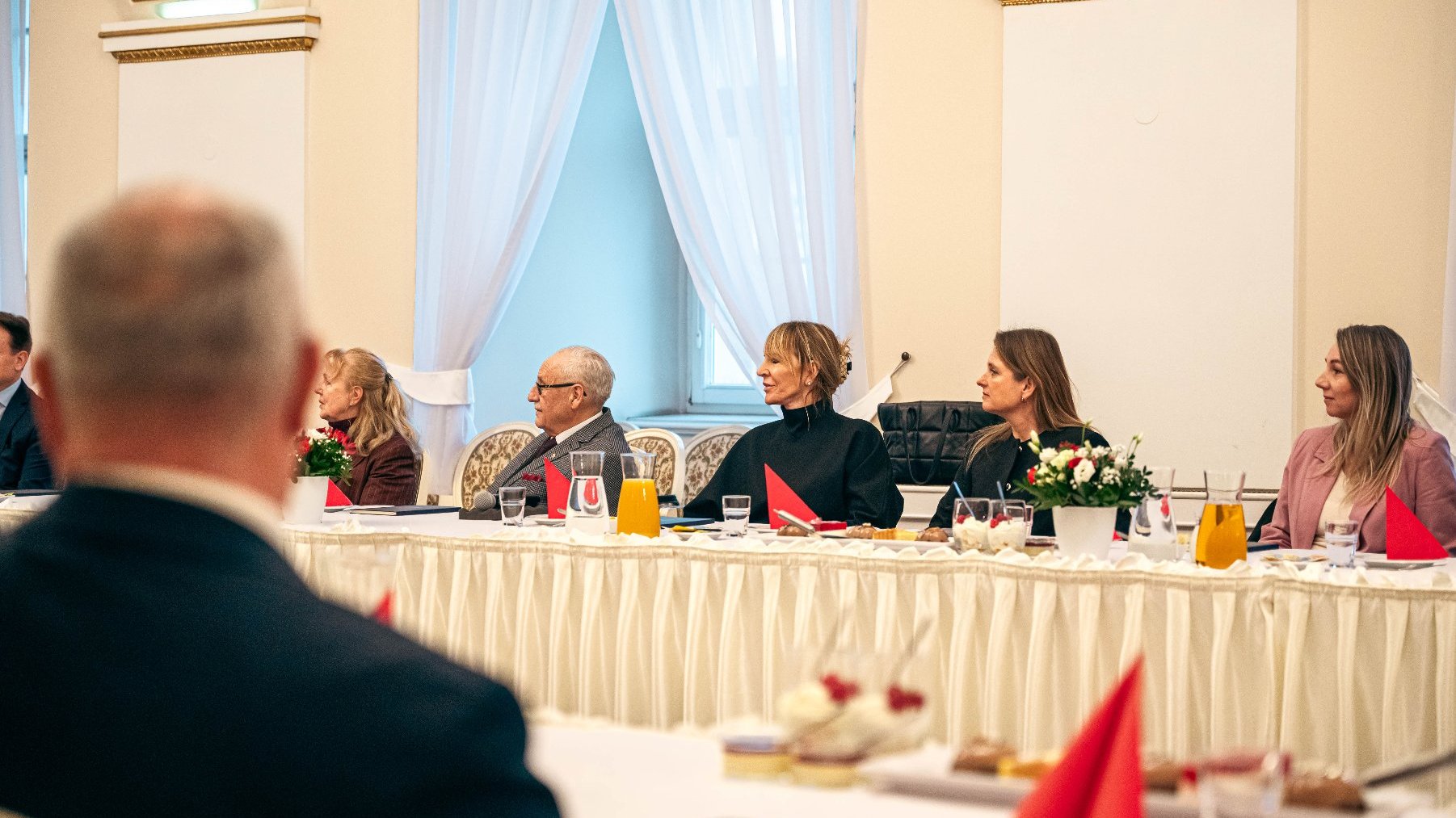 Zespół ludzi w formalnym spotkaniu w eleganckim pomieszczeniu. Stoł z białym fartuchem, czerwonymi ręcznikami, kwiatami i dzbankami z napojami. Osoby w formalnych strojach, rozmawiające lub słuchające prezentacji.