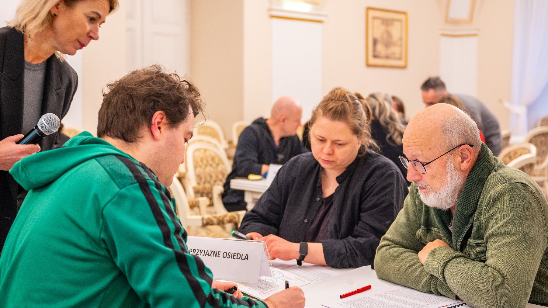 Na zdjęciu kilka osób przy stole, obok nich kobieta z mikrofonem w dłoni