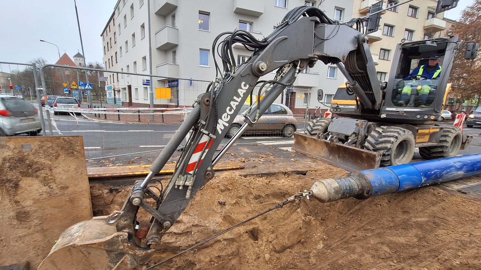 Zdjęcie przedstawia koparkę kopiącą dziurę w ziemi. Obok widać ulicę z samochodami i budynki.