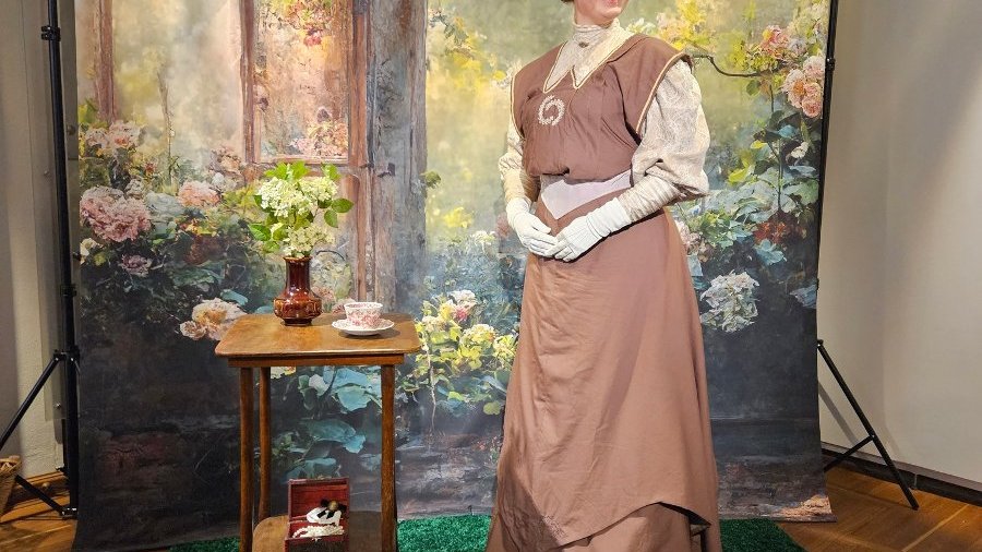 Kobieta ubrana w sukienkę stylizowaną na XIX wiek pozuje na tle fotograficznym. Sceneria przypomina ogródek.