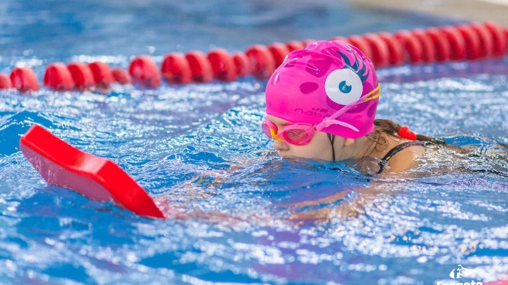 Dziewczynka w stroju kompielowym, w różowym czepku i goglach. Trzyma w rękach piankę i uczy się pływać w basenie sportowym.