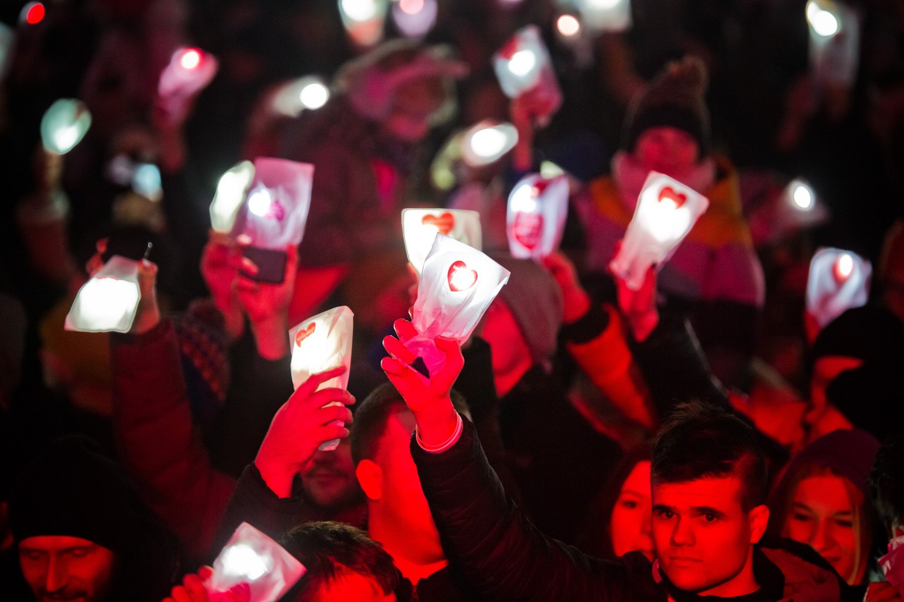 Ludzie ubrani w zimowe ubrania stoją w tłumie i wyciągają ręce do góry. W rękach trzymają telefony z zapaloną latarką, którą okrywa biała torba z naklejką serduszka. Torby przypominają lampiony. - grafika artykułu