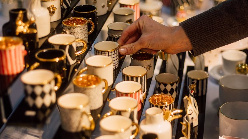 Kobieca ręka wyciąga rękę w stronę wystawki małych ceramicznych kubeczków, które są ozdobione w różne wzory.