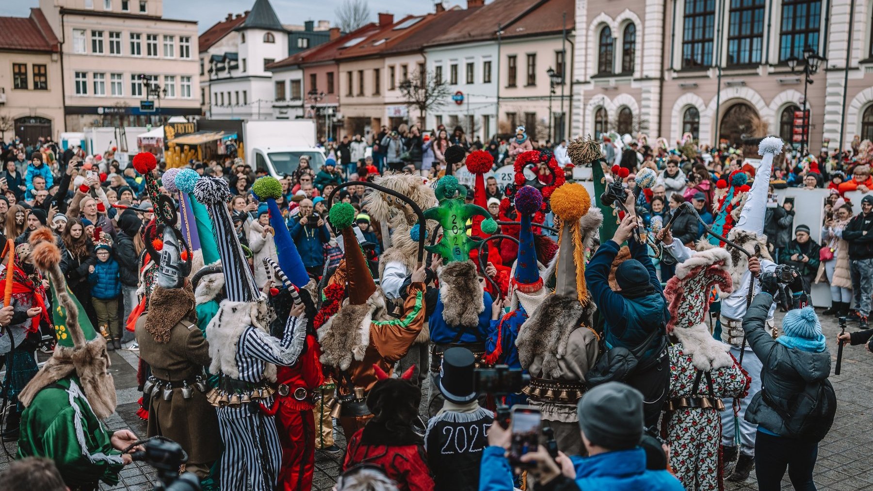 W centrum kadru znajdują się uczestnicy w różnorodnych, bogato zdobionych strojach ludowych. Wielu z nich nosi wysokie, spiczaste czapki udekorowane pomponami, futrzane elementy oraz pasy z dzwonkami. Część postaci ma maski, a niektórzy trzymają rekwizyty, takie jak kosy czy kije. Wokół nich zgromadzony jest tłum widzów, z których wielu robi zdjęcia i nagrywa filmy. W tle zabytkowe budynki z charakterystyczną architekturą, a także stragany i dekoracje.